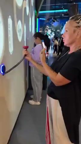 The video shows two women interacting with an interactive display on a wall in what appears to be a museum or exhibition. The woman in the foreground is closer to the camera. She has blonde hair in braids and is wearing a black t-shirt and white sweatpants with red stripes. She is holding a red handset to her ear with one hand and holding up her phone with the other, as if taking a photo or video of the exhibit. The woman in the background is wearing a purple shirt and grey sweatpants and is also holding a similar red handset to her ear.

The wall is covered with a screen that displays historical-looking images and text in what seems to be a timeline. The interactive element involves a red handset connected to the wall by a coiled cord. A blue light surrounds the place where the handset is attached to the wall. The area in the background is colorful and features geometric designs and other people walking around, suggesting it is part of the same exhibit.
