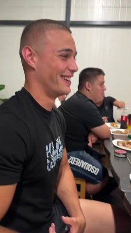 A group of young men are gathered around a table, appearing to be at a casual outdoor or semi-outdoor event. In the foreground, a man with a buzz cut and a black t-shirt is smiling and talking, gesturing with his hands. He is wearing black shorts with white lettering. Behind him, other men are seated at the table, some are also smiling and talking. The table is laden with food, plates, glasses, and numerous green bottles, some of which appear to be beer. There are also red bull cans and other beverages. The environment appears to be a covered patio or open-sided building with corrugated metal siding visible in the background. The lighting suggests it is daytime. The overall atmosphere seems relaxed and social.
