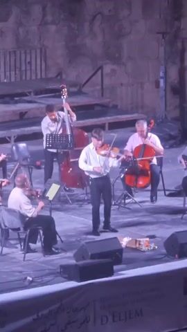 The video shows a string orchestra performing on a stage with a stone wall backdrop and wooden risers in the background. The lighting appears to be stage lighting with some purple hue. In the foreground, there is a stage with black speakers. The orchestra consists of several musicians playing string instruments. A young man in a white shirt and black pants stands in the center playing the violin. To his left, an older man sits and plays the violin. To the right of the central violinist, a man in a pink shirt sits and plays the cello. Behind the central violinist and to the left, a man stands and plays the double bass. Another musician, possibly playing the cello, is partially visible on the far right. There are music stands in front of most of the musicians. A small bouquet of flowers rests on the stage in front of the central violinist. A banner with Arabic script and the words "FESTIVAL INTERNATIONAL DE MUSIQUE SYMPHONIQUE D'EL JEM" is visible at the bottom edge of the frame.