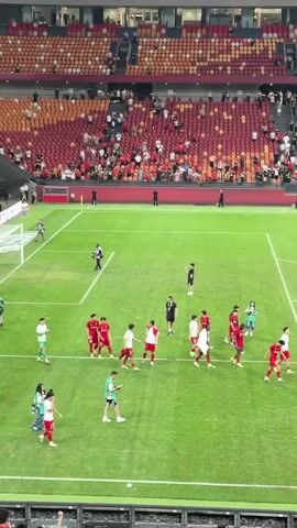 The video shows a soccer team walking off the field while their fans cheer from the stands. The soccer players, mostly wearing red jerseys, walk towards the edge of the field near the camera. Some are also wearing white or green jackets. The camera briefly shifts focus from the players on the field to the stands, where fans wearing black shirts raise their arms and cheer. The stands are colored in red, orange, and yellow patterns. Banners with Chinese writing and logos are displayed in front of the cheering fans.