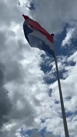 The video shows a close-up shot of a flag on a pole against a cloudy sky. The flag is red, white, and blue, and it is flapping in the wind. The sky is partly cloudy, with some dark clouds and some white clouds. The sun is shining through the clouds.
