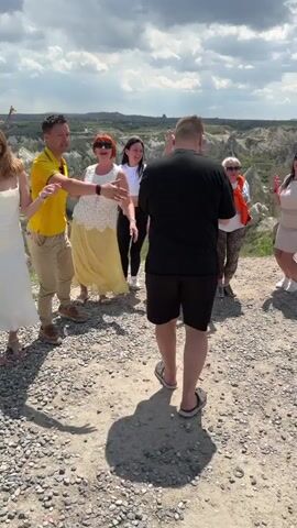 A group of approximately ten people is seen dancing on a gravel-covered area in Göreme, Turkey. The background features distinctive, light-colored geological formations under a partly cloudy sky with patches of blue. The individuals, dressed in casual summer attire including shorts, dresses, and t-shirts, are visibly enjoying themselves. They are smiling and laughing, with many raising their arms and moving energetically to what appears to be music. One person in a yellow shirt gestures and interacts with the group. The bright daylight suggests a warm, sunny day. No discernible text is visible on clothing or in the environment.