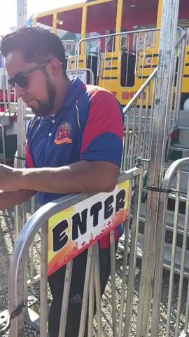 This media file captures a man in a blue and red polo shirt, sunglasses, and dark pants, standing at the entrance of an amusement park ride. He appears to be an attendant, as he is leaning on a metal railing with an "ENTER" sign. The sign, in a bold, colorful font, features a silhouette of a city skyline and a sunset. Behind him, a yellow and red amusement ride with the words "CRAZY BUS" visible on its side is partially obscured by the railing and the attendant. The ride has multiple windows and appears to be a bus-themed attraction.

In the background, a young child with curly blond hair, wearing a white shirt, is visible, hinting at a family-friendly atmosphere. The attendant is holding what appears to be a phone in one hand and gesturing with the other, possibly interacting with a guest or checking something. A sign on the ride indicates "THIS RIDE 28 CREDITS" and lists safety information, suggesting an operational amusement park. The bright sunlight and clear sky suggest it's daytime, likely a sunny day, and the overall scene evokes a sense of fun and activity associated with an amusement park. The location context of Gloucester, United States of America, further places this scene within a typical American amusement or fairground setting.