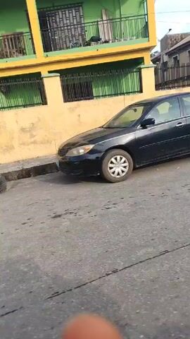 The video shows a street in what appears to be a residential or commercial area in Nigeria. The camera moves slowly, showing a parked car with damage to the rear, a man walking across the street, a street vendor, a woman walking by, a person kneeling, and a car and motorcycle driving. The buildings are varied in style and condition, with some appearing older and others more modern. The sky is partly cloudy.