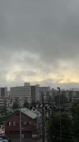 This media file captures an urban scene in Yachiyo, Japan, under a heavy, overcast sky. The image is dominated by a dense formation of clouds, which obscure most of the sky, creating a somber atmosphere. Hints of yellowish light, possibly from the setting or rising sun, can be seen peeking through gaps in the clouds towards the right side of the frame.

In the midground, a cluster of multi-story apartment buildings forms the core of the urban landscape. These buildings are a mix of white and muted tones, with many balconies visible. To the left of these larger buildings, a smaller, reddish-brown, two-story structure with a peaked roof stands out. Its windows are dark, and there appears to be some red text on its facade, though it is not clearly legible.

In the foreground, a network of utility poles and wires crisscrosses the frame, adding a layer of urban infrastructure to the scene. Some of the poles have speakers mounted on them, which are common in Japanese residential areas for public announcements. Dense green trees are visible on the right side, partially obscuring the apartment buildings and adding a touch of nature to the otherwise built environment.

A street is visible in the lower left corner, with several parked cars. The overall impression is one of a quiet, possibly residential area, on a cloudy day. The weather appears to be either approaching rain or has recently passed. The time of day is difficult to pinpoint precisely due to the cloud cover, but the quality of light suggests either early morning or late afternoon. The mood conveyed is one of subdued calm, perhaps a touch melancholic due to the cloudy sky. There are no people visible in the frame.