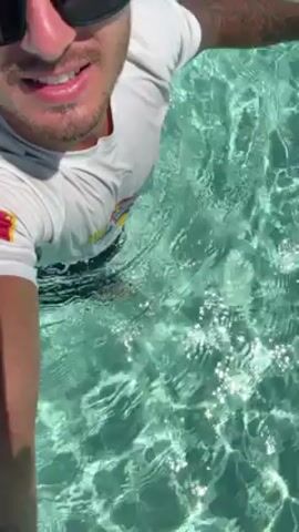 A man is standing in shallow, clear water. He is wearing a white t-shirt and sunglasses, and is smiling. The water is turquoise and ripples in the sunlight.