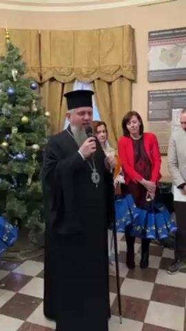 A man in a black religious robe is speaking into a microphone. He has a long beard, a black hat, and a necklace with a cross. He is holding a long staff in his left hand. Standing behind him are three other people. To his right is a woman in a red jacket, holding several gift bags. To her left is another woman in a traditional embroidered shirt. Further to the right of the first man stands another man in a grey blazer and jeans. Behind them are walls with posters on them. To the left of the first man is a Christmas tree decorated with ornaments. The floor is tiled in a black and white checkerboard pattern.