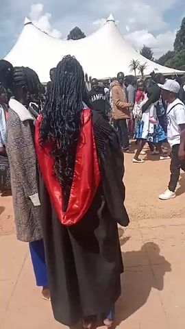 The video shows a crowd of people, many wearing graduation gowns, gathered on a brick-paved area outdoors under a partially cloudy sky. A large white tent is visible in the background. People are walking around, and some appear to be talking to each other. There is a banner with an emblem and lettering visible on the left side. Debris such as an empty plastic water bottle can be seen on the ground.  The camera angle is slightly low, following a woman with braided hair wearing a graduation gown with a red hood.