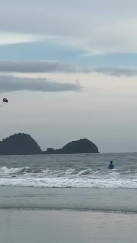 A drone shot pans across a beach with gentle waves. In the distance, two lush green islands rise from the sea. In the sky, a parachute with a person attached drifts lazily. Closer to shore, several people are wading in the water, and one person is surfing on a board. The sky is partly cloudy with a mix of gray and blue. The overall mood is serene and relaxed.