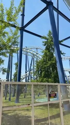 A roller coaster train, carrying several passengers, navigates a curved section of track against a clear blue sky. The roller coaster structure, featuring large blue support beams and white track, is prominently visible. A lush green tree stands behind the track, partially obscuring the view. In the foreground, a metal fence with a caution sign that reads "DANGER PERICOLO DI MORTE VIETATO ENTRARE" (Danger Death Danger Forbidden Entry) is present. The ground appears to be covered in grass and some water features are visible in the background.