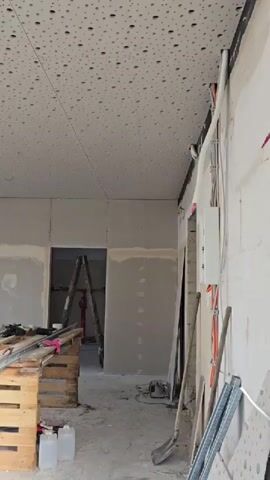 This media file depicts an interior construction site in Haren (Ems), Germany. The scene is an unfinished room with a perforated ceiling and bare drywall. Various construction materials and tools are scattered throughout the space, including wooden pallets piled with planks, a ladder, buckets, bags of construction material, and electrical conduits. Sunlight streams in from large windows, suggesting it is daytime, and the weather appears fair.

The overall impression is one of active construction or renovation. The presence of materials like drywall, bags of what appears to be cement or plaster, and tools indicates that work is in progress. The room itself appears to be a commercial or residential space undergoing significant transformation. The exterior view through the windows shows a street with residential buildings and parked cars, consistent with the provided location context of Haren (Ems), Germany. No people are visible in the frame, focusing the attention on the environment and the ongoing work. The lighting suggests a bright, possibly sunny day.