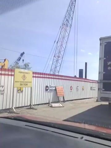 From a vehicle, the camera captures a construction site under a bright blue sky. A large crane is visible to the left. A white fence with a red stripe runs along the perimeter, displaying warnings and company logos, including "Danantara Indonesia." Orange traffic barriers are placed along the base of the fence. A large truck passes by on the right. The construction site includes large metal beams being lifted into place by the crane.