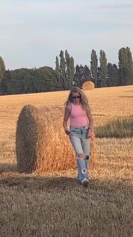 A woman with blonde curly hair wearing a pink tank top and ripped light blue jeans stands in a dry field with hay bales. She is holding a phone in her hands and appears to be looking at it. The sun is low in the sky, casting a warm glow over the scene. The field is surrounded by trees.
