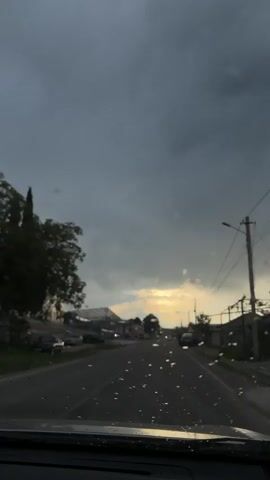The video shows a view from inside a car looking out onto a wet road. The sky is overcast with a hint of sunset peeking through the clouds in the distance. There are buildings and parked cars on either side of the road, as well as utility poles with wires. The windshield has raindrops on it, and there is music playing in the background.