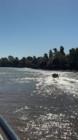 A small motorboat containing several people is moving across a wide body of water, leaving a wake behind it. The water is brown, reflecting sunlight, creating a shimmering effect. The shoreline in the background is lined with trees and houses. The sky is clear and blue. A metal railing is visible in the foreground.