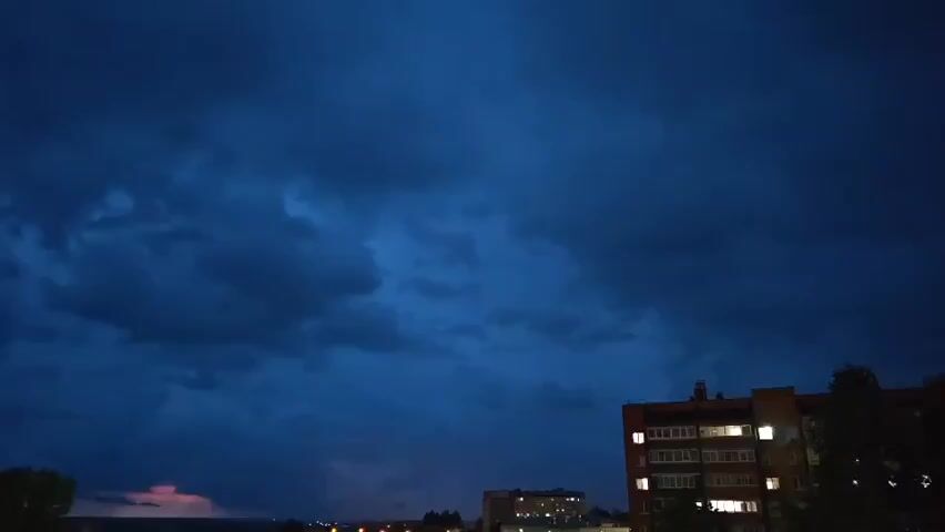 A nighttime cityscape under a dark, cloudy sky is shown. On the right side of the frame, a brick apartment building with lit windows stands tall. In the distance, more buildings and lights of the city are visible. There is a faint pinkish hue in the sky to the left, possibly indicating distant lightning or sunset. The overall mood is dark and atmospheric, suggesting an approaching storm or the aftermath of one.
