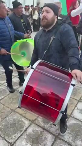 The video shows a group of people performing music outdoors, seemingly during a festive event. There are four musicians visible in the foreground: one playing a red, cylindrical bass drum, another with a fluorescent green drum or tambourine-like instrument, a clarinetist, and someone playing what appears to be a bagpipe. The drummer in the foreground is a man with a beard, wearing a black beanie and a grey patterned sweatshirt. He's striking the red drum with a stick in each hand. The person with the bagpipe is holding a Bulgarian flag. Several other people are visible in the background, some in traditional clothing, and some onlookers. The ground is paved. It appears to be a daytime outdoor event in a village or small town, possibly a cultural celebration.