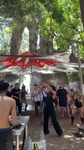 Here's a detailed description of the video:

**Scene:** The video captures an outdoor party or gathering under the shade of large trees and a makeshift canopy.

**Environment:** The scene is set in a forested or wooded area, likely during the day. The canopy above the party area appears to be constructed from fabric, with a geometric or patterned design. Mist or vapor is being dispersed in the air, creating a haze effect and potentially helping to cool the area.

**People:** A group of people are present, dancing and enjoying the music. There is a mix of genders and ages. Some of the individuals are wearing casual clothing. One person is shirtless and seemingly acting as a DJ, operating equipment on a folding table.

**Objects:** Key objects include:
*   DJ equipment (mixers, turntables, or similar devices).
*   A folding table holding the DJ equipment.
*   Bottles and cans on the table, suggesting drinks.
*   A makeshift fabric canopy, providing shade.
*   Large trees surrounding the area.
*   A sign to the right of the canopy.

**Actions:** The primary action is dancing. People are moving to the rhythm of the music. The person behind the DJ equipment is manipulating the controls, suggesting they are mixing or playing music.

**Interactions:** People are interacting through dancing and enjoying the shared atmosphere. The DJ is providing the music that motivates the movement and energy of the crowd.