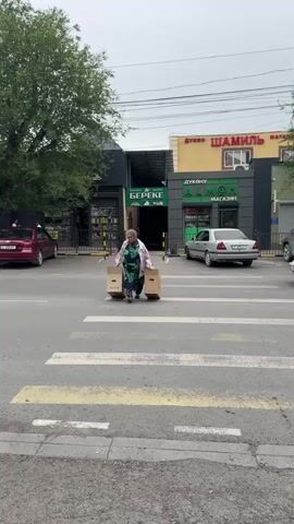 A woman in a green and black patterned dress and a pink jacket walks across a crosswalk carrying two cardboard boxes. Cars are stopped on the street, and a yellow van is driving away. Buildings with storefronts are visible in the background.