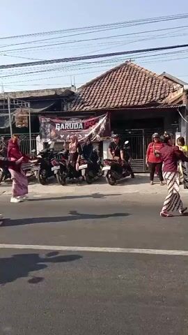 A group of people, some in traditional clothing, are marching down a street. The people in traditional clothing appear to be performing some sort of dance or procession. Several motorcycles are parked along the side of the street, and some people are watching the procession from the sidewalk. The buildings in the background appear to be shops or small businesses. The sky is clear and the sun is shining.