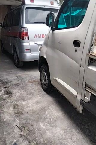The video shows the side of a white van with the word AMBULANCE visible on the back of a silver vehicle parked behind it. A round sign with a red P symbol on a white background and a black border is attached to the white van. The ground appears to be concrete and is dirty. The video is filmed from a low angle and is rotated.
