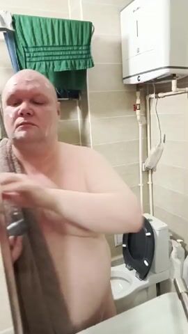 A bald man wearing a brown towel around his shoulders is applying white cream to his face in a bathroom. He holds a black tube of product in his left hand and uses his right hand to spread the cream on his cheeks, forehead, and chin. The bathroom has tiled walls, a green towel hanging on a rack, a mirror reflecting the scene, a white electric water heater on the wall, and a toilet seat visible in the background.