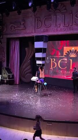 A magician performs a balancing act on a stage. They are upside down, balanced on a chair with their legs extended upwards. A tall, stacked tower of black and white boxes is precariously balanced on their feet. Confetti is scattered on the stage around the performer and their equipment. Two assistants stand on either side of the stage. The background features a large screen with the words "BELLIS DELUXE HOTEL" displayed. The stage is lit with purple and white lights. The audience is visible in the foreground.