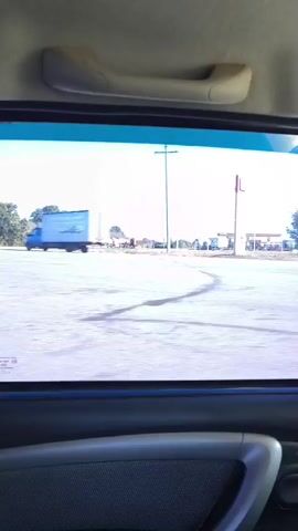 The video shows a view from inside a vehicle, looking out the rear window. The window is tinted blue at the top. Outside, a road is visible, with a few trees and some buildings in the distance. There are also utility poles and some vehicles on the side of the road. The weather appears to be clear and sunny. The video is shot from a moving vehicle, as evidenced by the slight blur and the changing perspective. There are no people visible in the video.
