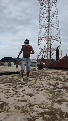 A shirtless young man wearing white goggles and denim shorts stands on a rooftop. He is in the foreground, facing forward with his arms at his sides. Behind him, a tall, red and white lattice tower dominates the background, likely a cell tower. To the left of the tower, there are buildings with satellite dishes, and further left, a dark-roofed house. To the right of the tower, there's a glimpse of trees and a rusty, conical structure.

The sky is overcast with a grayish hue, suggesting cloudy or possibly pre-rain weather. The time of day appears to be daytime, given the ambient light. The rooftop surface is a textured, light-colored concrete. The man's stance and the slight turn of his head suggest he might be posing or in the midst of an activity, possibly dancing or preparing to move. The overall mood is somewhat casual and urban. No visible text is present in the frame. The scene is set outdoors in what appears to be a residential or semi-industrial area.