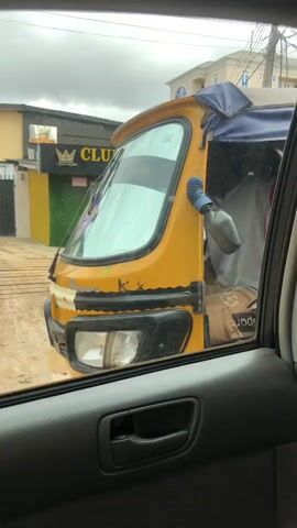 A yellow auto rickshaw with a blue and white canopy is parked on a dirt road. The driver, wearing a grey sleeveless shirt and shorts, is seated in the driver's seat. A person in black is visible in the passenger area of the auto rickshaw. To the left of the auto rickshaw is a building with a green and yellow facade and a sign that reads "CLUB HOXEGEN GBAGADA". Two people, a man and a woman, are standing near the entrance of the club. A series of wooden planks are laid across the dirt road in front of the club. The scene is viewed from inside a vehicle, looking out the window. The sky is overcast.