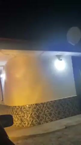 A car approaches a building at night. The building has a tiled base and a white exterior. There are exterior lights. The building has an arched entryway and a window. There is a gate visible in the entryway. The camera is inside the car.