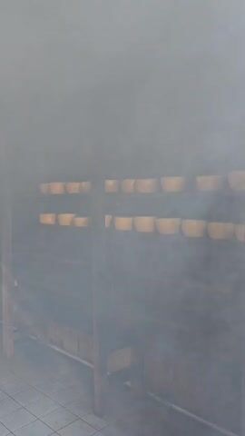 The video shows a hazy indoor space, possibly a smoking room, with shelves lined with rows of what appear to be round cheese blocks. Sunlight streams in through openings, highlighting the smoky atmosphere. A metal chimney and wooden support beams are visible. The floor appears to be tiled.