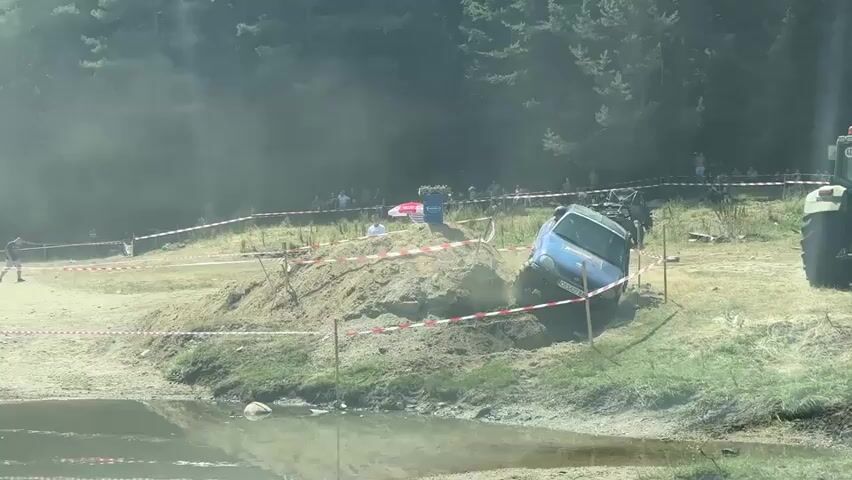 A blue off-road vehicle is stuck on a dirt embankment next to a body of water. The vehicle is angled significantly, with its front wheels appearing to be lower than its rear wheels. Dust or smoke is billowing around the vehicle, suggesting it is either stuck or has just encountered an obstacle. Red and white caution tape is visible, marking off an area around the embankment. Several people are standing in the background, observing the scene. A tractor is partially visible on the right side of the frame. The environment appears to be a wooded area, with trees in the background. The sun is shining brightly, casting light and creating some glare and haze in the image.