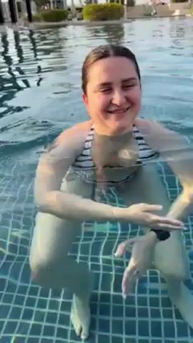 A young woman is sitting in a swimming pool with light blue water, smiling at the camera. She has dark hair and fair skin, and is wearing a black and white striped bikini top and a black smartwatch on her left wrist. She is sitting on the tiled bottom of the pool, with her legs spread apart. Her hands are visible under the water, moving slightly. The pool has small square blue-green tiles. The background includes a blurry view of trees and some kind of structure.
