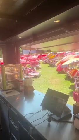 The video pans across a sun-drenched outdoor seating area with numerous red and yellow striped beach umbrellas and lounge chairs. The scene appears to be a resort or pool area. In the foreground, a metallic counter is visible with a cash register or payment terminal. To the left of the counter, a glass display case with illuminated shelves contains food items. The background shows a lush green lawn dotted with people relaxing under the umbrellas. The lighting suggests it is daytime.