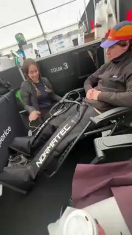Two individuals are seated and equipped with Normatec leg compression devices. The person on the left is a woman with long hair in braids, wearing a dark jacket. She is seated in a black chair, and her hands are near the device. The person on the right, also seated in a black chair, is wearing a dark jacket and a colorful baseball cap. The environment appears to be a tent or temporary structure, with black staging and other equipment visible in the background. A cup is visible in the foreground.