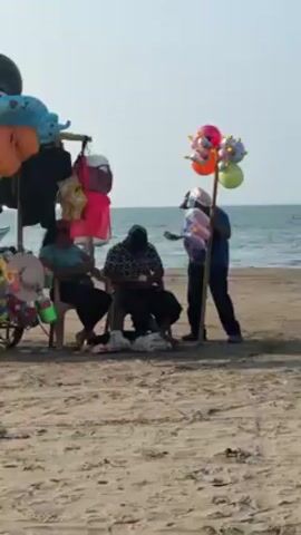 The video shows a beach scene on a bright day. In the foreground, a man with a baseball cap is selling balloons attached to a wooden stick. The balloons are various colors (pink, orange, green, blue) and some have small yellow rubber ducks attached to them. The man is standing in front of a seated group of people by a rack filled with other merchandise, including stuffed animals and bags. In the background is the ocean with calm waves under a clear sky.