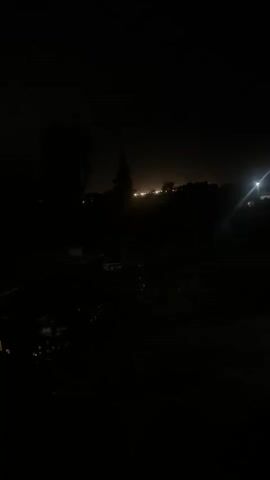 The video shows a dark, nighttime outdoor scene. The foreground is mostly black with some indistinct shapes suggesting foliage. In the middle ground, there are silhouettes of trees and possibly some low buildings. In the background, there is a faint glow of lights from a town or city on the horizon. There is also a bright, artificial light source in the middle right of the frame, creating a lens flare. The overall impression is one of a quiet, rural or suburban night with distant urban illumination.