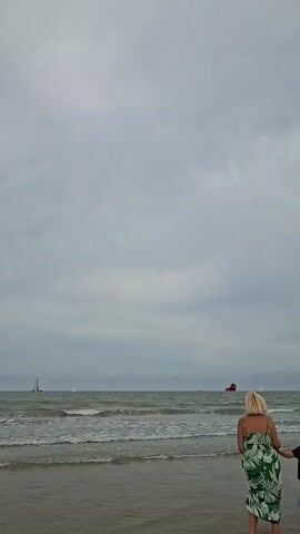 A woman with blonde hair and a green floral dress stands on a beach with her back to the camera, holding hands with a child whose face is not visible. The woman is wearing sunglasses. In the background, the ocean waves crash onto the shore under a cloudy sky. Several sailboats and a red cargo ship are visible on the horizon. The camera is focused on the people and the sea.