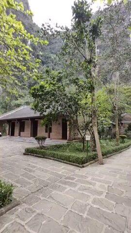 The video shows a stone-paved pathway leading towards a building in a natural setting. The pathway is flanked by trees and manicured hedges. In the background, a mountain rises, partially obscured by clouds. The building in the foreground appears to be constructed of brick, with a dark roof and multiple doorways. Further down the path, a second, smaller building is visible. A few people are walking along the pathway in the distance. The overall scene suggests a peaceful and well-maintained outdoor space, possibly a park or garden within a mountainous region.