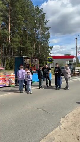 The video shows a scene at a roadside fair or market, with several food and game stands set up along the edge of a road. People of various ages are gathered around the stands. There's a game booth with stuffed animals, a hot dog stand, and other vendor setups. Some individuals are interacting with the vendors, while others walk by or stand in small groups. A man pushes a stroller. Bicycles are also visible. The background features a mix of trees, buildings, and a cloudy sky.