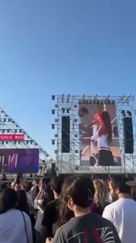 The video shows a crowd of people at an outdoor music festival. In the background is a stage with large screens displaying a performer. The festival appears to be the "Italia Music Fest." The sky is clear and blue, suggesting it is daytime. The camera focuses on various individuals in the crowd, showing their expressions and movements. The audience seems engaged and enjoying the performance. At one point, the camera abruptly focuses on a young man's face, showing a close-up with an exaggerated expression.