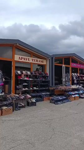 A street view shows several small shops lining a paved area. The shops have large glass fronts and are stocked with shoes displayed on shelves and on the ground. Some of the shops have signs above their entrances. The sky is cloudy. A few cars and trees are visible in the background. The overall scene appears to be an outdoor market.