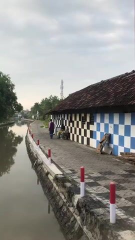 The video shows an outdoor scene next to a body of water. On the right, there is a building with a checkered facade of blue and white tiles and a dark, tiled roof. Next to the building is a paved walkway with a checkered pattern. Red and white posts line the edge of the walkway. A person wearing a purple jacket and dark pants is walking on the walkway. Further down the walkway, another person is seated. The body of water, possibly a canal, is on the left side of the frame, with a stone wall along its edge. Trees and greenery are visible in the background. The sky is cloudy.