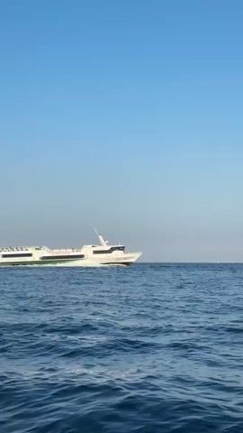A wide shot captures a white passenger ferry with green accents on its side, sailing across a deep blue, moderately choppy sea. The sky above is a clear, cloudless blue. The ferry is moving from left to right across the frame, leaving a white wake behind it. The image is framed by the edge of a wooden structure, likely part of another boat or dock, in the lower right corner. The scene appears to be filmed from another vessel.