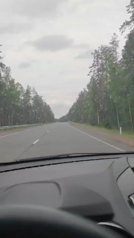 The video shows a point-of-view shot from inside a car driving on a two-lane road. The road is surrounded by a dense forest of pine trees on both sides. The sky is overcast with clouds. There are raindrops visible on the windshield, suggesting it is raining or has recently rained. The car is moving forward, and the road stretches into the distance, with a white line marking the lane. The dashboard of the car is partially visible in the foreground.