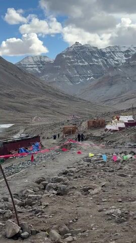 The video shows a mountainous landscape with a rocky foreground. In the distance, there are tall mountains covered in snow. The sky is partly cloudy with blue patches. There are small buildings and structures in the mid-ground, along with colorful flags and pink caution tape marking off an area. Several people are walking along a path towards the mountains. The scene appears to be a remote and rugged location.