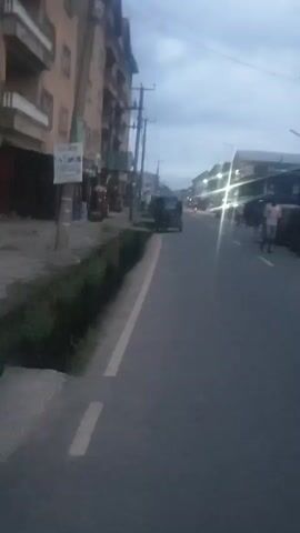 The video shows a street in what appears to be a developing or urban area. It is either dusk or dawn given the low light.  The road is asphalt and has a yellow line marking the edge. There's a low hedge or planting along the side of the road. On one side of the street there is a multi-story building and on the other side there are single-story structures that appear to be shops or businesses. There are people and vehicles including auto rickshaws visible on the road. The video is somewhat blurry, which may affect visibility.