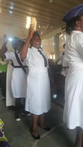 In this video, a group of women, likely members of a uniformed organization such as a church choir or youth group, are participating in what appears to be a religious service or celebration. They are dressed in white uniforms with ties and caps, suggesting a formal occasion. The setting is indoors, possibly a church hall or community center, with a wooden ceiling and exposed beams. The lighting is bright, indicating it could be daytime.

The woman in the foreground is the main focus, with her arm raised and mouth open as if singing or speaking with enthusiasm. Her expression is joyful. Other women in the background are also engaged, with some raising their hands and smiling. The overall atmosphere seems to be one of shared joy and spiritual engagement.

The video is short, lasting only a few seconds, and the audio seems to contain singing or chanting in what could be a Nigerian language, given the location context. The image quality is moderate, with some blurriness, suggesting it was recorded with a mobile phone. There is no visible text in the video. The presence of chairs and the general decor suggests a communal gathering.
