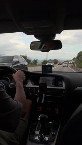 From the interior of a car, the driver’s hands are on the steering wheel, driving on a two-lane highway. Cars, including a truck, are visible on the road ahead. The driver is wearing a gray shirt and shorts. A mobile phone is mounted on the dashboard. A small Kazakh flag is hanging from the rearview mirror, and a picture of a baby is affixed to the mirror. The driver's reflection can be seen in the mirror. The surroundings include mountains and trees. The sky is partly cloudy.